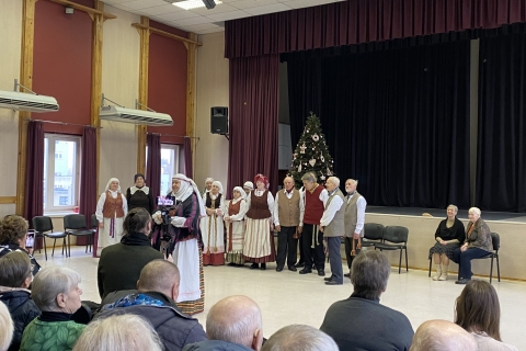 Grupė žmonių tautiniais drabužiais yra šalia scenos prie kalėdinės eglutės