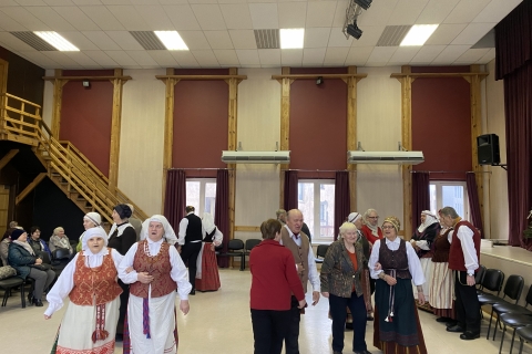 grupė žmonių didelėje salėje. Dalis jų vilki tradicinius liaudies kostiumus. Jie stovi poromis, ruošiasi šokti. Kiti sėdi šone ir stebi renginį.