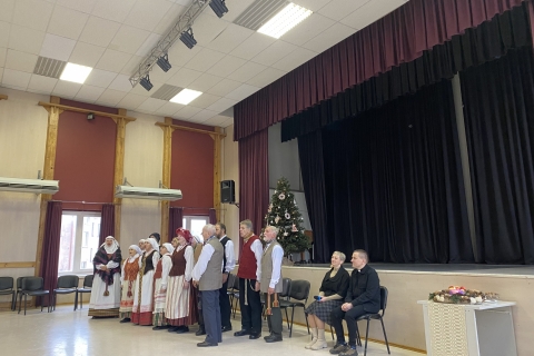 grupė žmonių su tradiciniais drabužiais salėje, šalia jų sėdi du žmonės. Fone scena su užuolaidomis ir Kalėdų eglute, priekyje stalas su dekoracijomis ir žvakėmis.