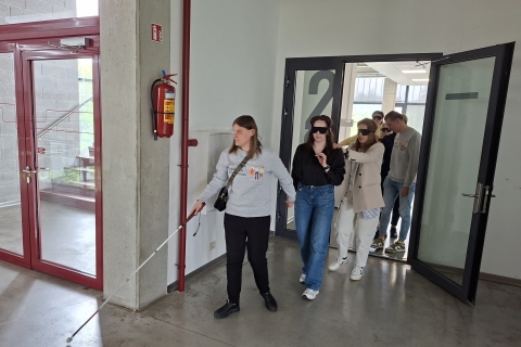 Grupė žmonių eina koridoriumi. Priekyje moteris su baltąja lazdele veda kitus, ji laiko už rankos kitą žmogų. Visi už jos einantys dėvi a