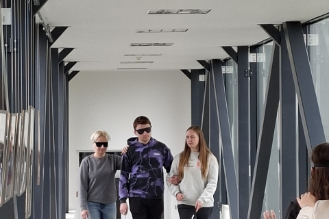 Keturi žmonės moderniame koridoriuje. Du iš jų uždengtomis akimis – vienas eina su baltąja lazdele, kitą veda už rankos. Juos lydi da