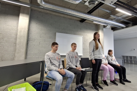 Keturi jauni žmonės modernioje auditorijoje. Trys iš jų (du vaikinai ir viena mergina) sėdi ant kėdžių, o kita mergina stovi šalia jų, pr