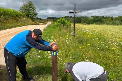 Du vyrai pakelėje įrengia medinį maršruto stulpą. Vienas jį prilaiko, kitas pasilenkęs dirba prie stulpo apačios_ ant stulpo pritvirtintas mėlynas ženklas su geltona rodykle