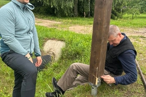 Du vyrai prie kaimo keliuko tvirtina aukštą medinį maršruto stulpą. Vienas sėdi ant žemės ir dirba prie stulpo apačios, kitas stovi šalia
