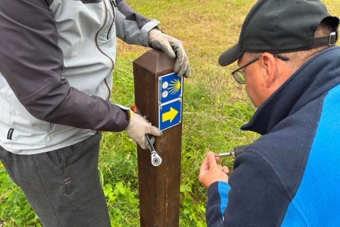 Du vyrai tvirtina Camino Lituano ženklus prie aukšto medinio stulpo. Vienas laiko stulpą ir veržliaraktį, kitas atsuktuvu tvirtina mėlyną lentelę su geltona kriaukle