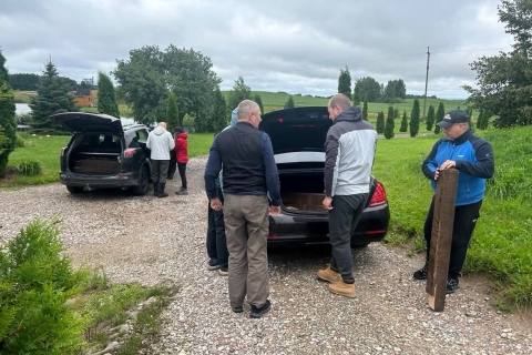 Keli žmonės prie automobilių ruošiasi darbams lauke. Atidarytos automobilių bagažinės, o dešinėje stovintis vyras rankose laiko ilgą medinį žymėjimo stulpą