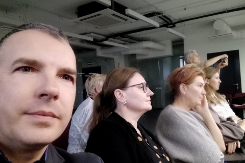 Žmonės sėdi patalpoje, panašioje į konferencijų salę. Atrodo, jog jie atidžiai kažko klausosi