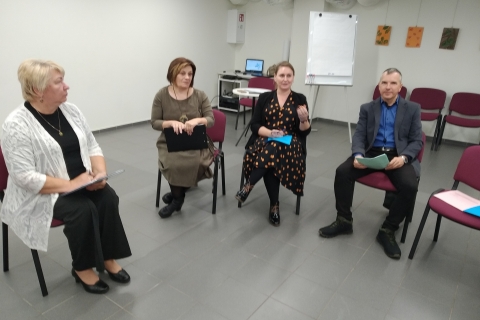 Puslankiu sėdi trys moterys ir vienas vyras. Vieni laiko sektuvus, o kiti - lapus_ Kambaryje taip pat matyti kompiuteris ir keli meno kūriniai