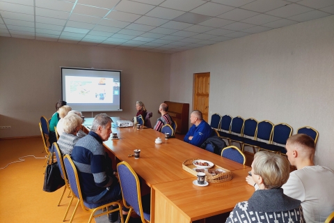 Žmonės sėdi prie stalo, stebi skaidrių projekciją ant sienos
