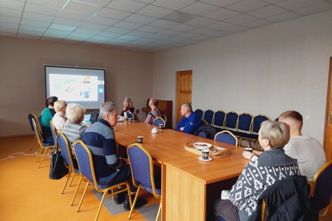 Prie stalo sėdi grupė žmonių, ant stalo matyti puodelių, nešiojamas kompiuteris