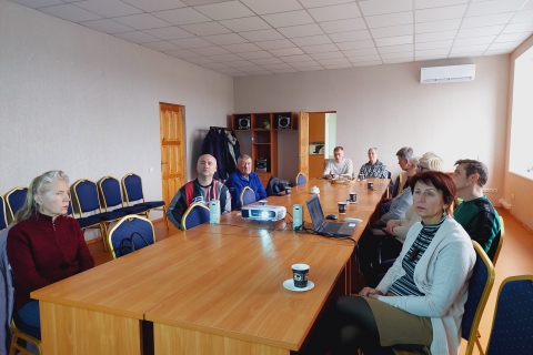 devyni žmonės susitikimų kambaryje sėdi aplink stalą su nešiojamu kompiuteriu ir projektoriumi. Kambarys šviesus, apšviestas natūralia šviesa